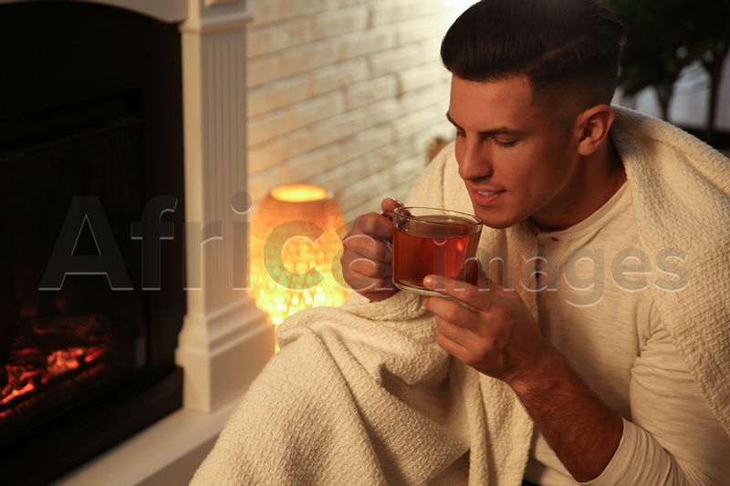 Man covered with white plaid enjoying cup of tea near fireplace