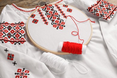 Photo of Shirt with red embroidery design in hoop, needle and thread on table, closeup. National Ukrainian clothes