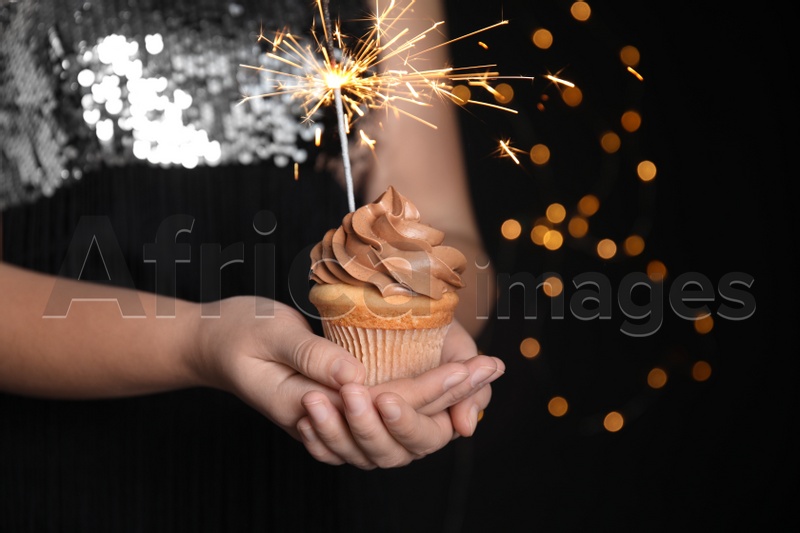 Photo of Woman holding birthday cupcake with sparkler on blurred background, closeup