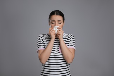 Sick young woman sneezing in tissue on grey background. Cold symptoms