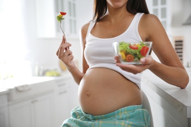 Young pregnant woman with bowl of vegetable salad at table in kitchen, closeup. Taking care of baby health