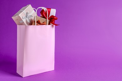 Photo of Pink paper shopping bag full of gift boxes on purple background. Space for text