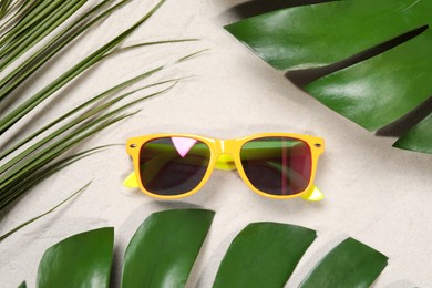 Photo of Stylish sunglasses and tropical leaves on sand, flat lay