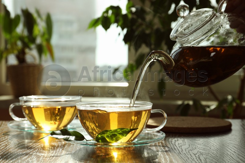 Photo of Pouring green tea into glass cup with saucer and leaves on wooden table