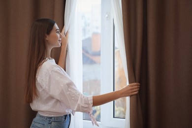 Woman opening elegant window curtains in room