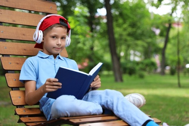 Photo of Cute little boy with headphones reading book on lounger in park