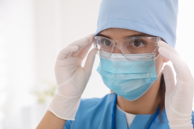 Doctor in medical gloves and protective mask putting on glasses against light background, closeup