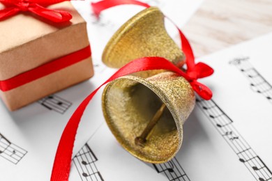 Photo of Golden shiny bells with red bow, gift box and music sheets on table, closeup. Christmas decoration
