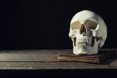 Photo of Human skull and old book on wooden table against black background, space for text