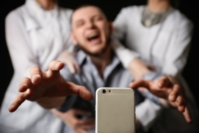 Emotional man reaching for smartphone while medical workers holding him back on black background, closeup. Addiction concept