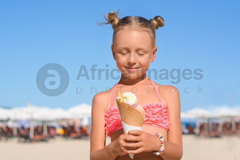 Adorable little girl in swimsuit with delicious ice cream at beach on sunny summer day