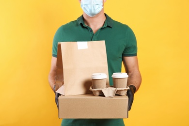 Courier in medical mask holding packages with takeaway food and drinks on yellow background, closeup. Delivery service during quarantine due to Covid-19 outbreak